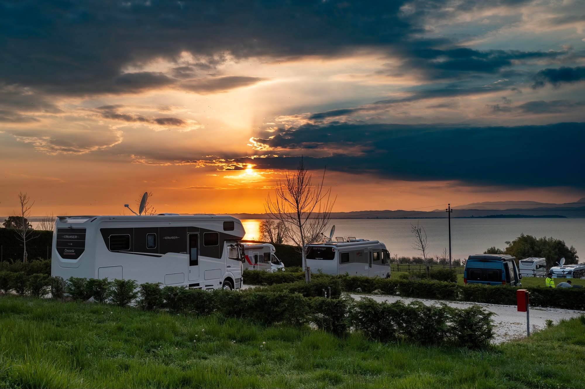 Wohnmobile auf einem Campingplatz mit Blick auf einen See bei Sonnenuntergang unter einem bewölkten Himmel.