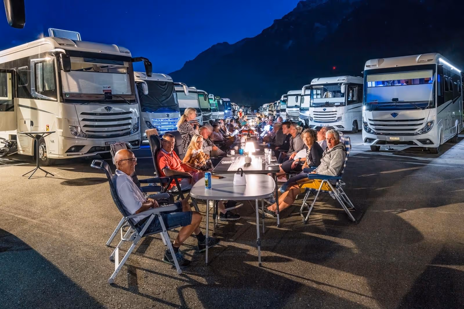 Gruppen von Menschen sitzen abends an langen Tischen im Freien zwischen Reihen von Wohnmobilen mit beleuchteten Lampen.