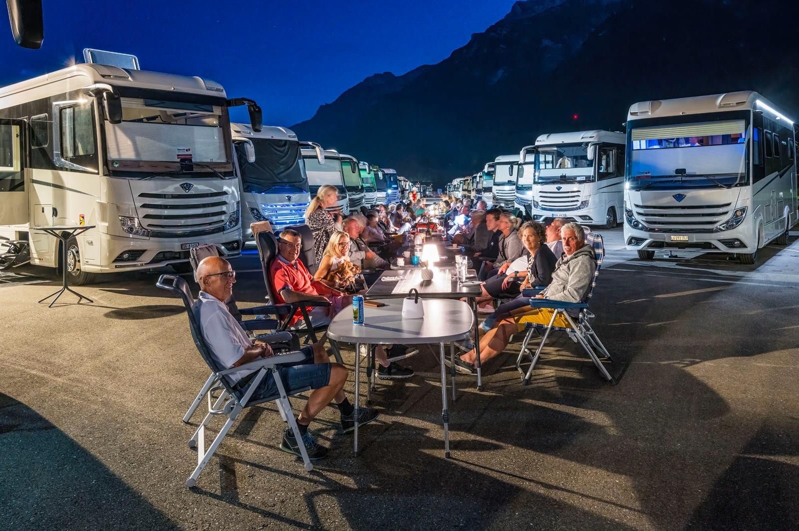 Gruppen von Menschen sitzen abends an langen Tischen im Freien zwischen Reihen von Wohnmobilen mit beleuchteten Lampen.