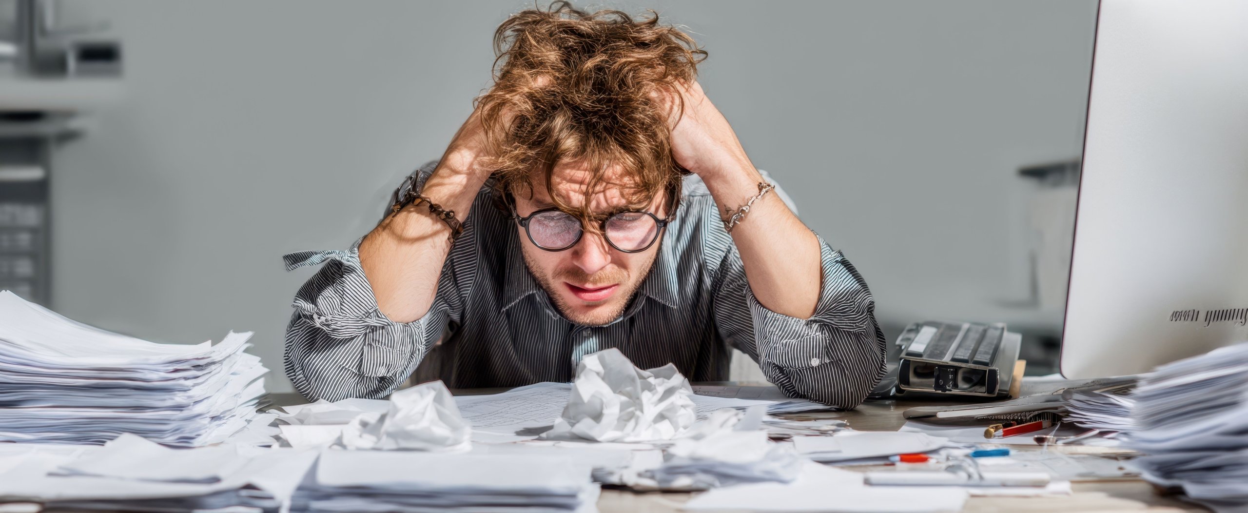 Gestresster Mann mit zerzausten Haaren und Brille am Schreibtisch, umgeben von Stapeln und zerknülltem Papier.