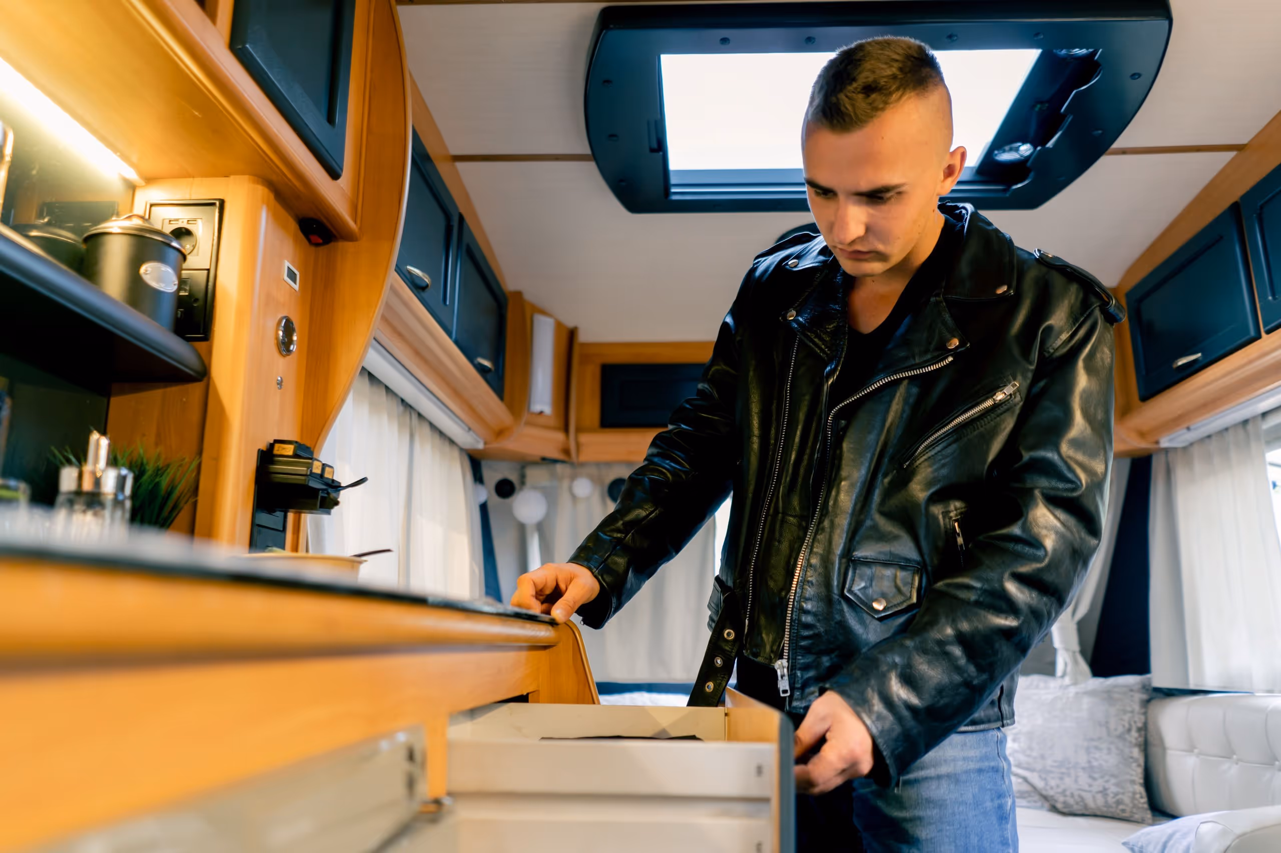 Ein junger Mann in schwarzer Lederjacke öffnet eine Schublade in einem Holzinterieur eines Wohnmobils.