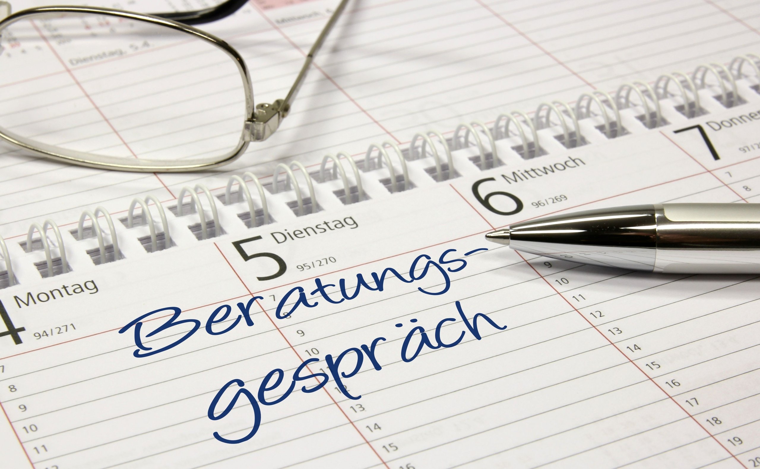 Kalender zeigt die Tage Montag den 4., Dienstag den 5. und Mittwoch den 6. mit eingetragenem handgeschriebenem Text Beratungs-gespräch, daneben liegt ein silberner Kugelschreiber und eine Brille.