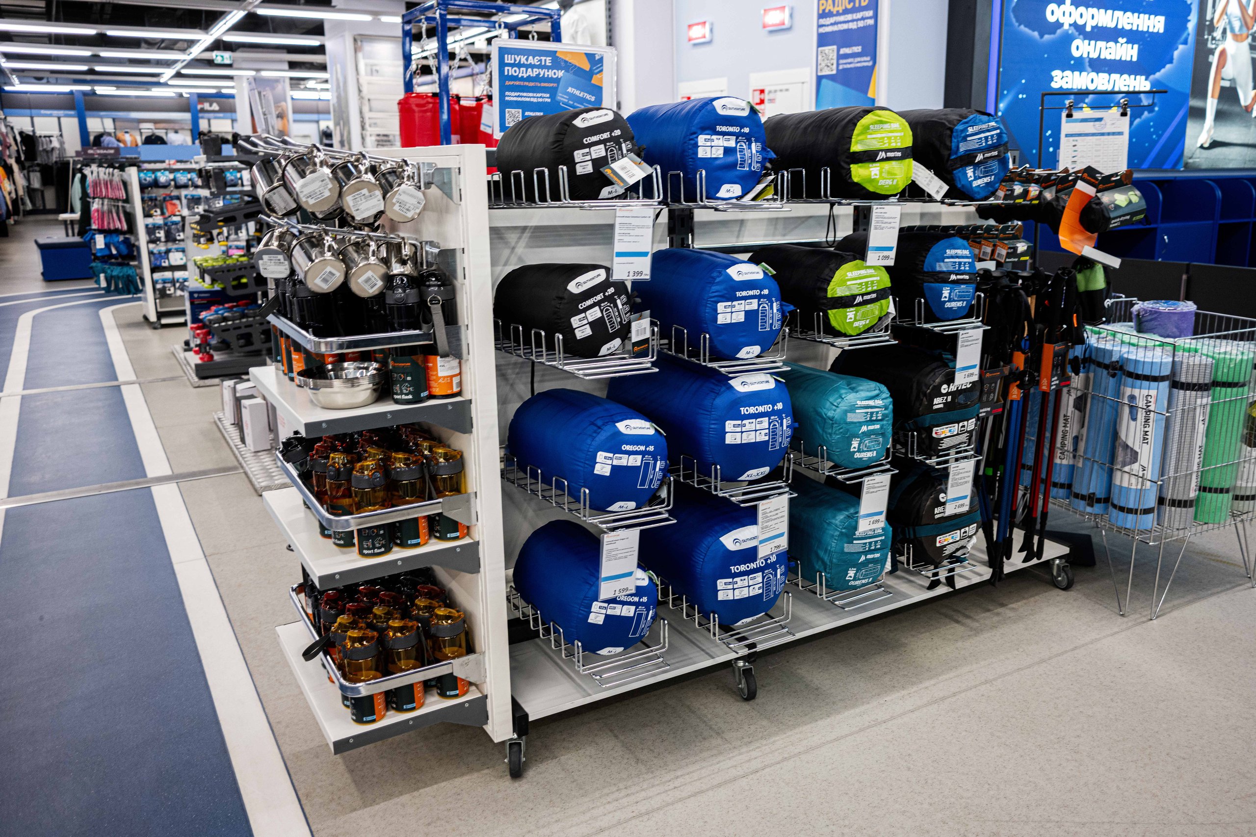 Regal mit zusammengerollten Schlafsäcken, Wasserflaschen und Wanderstöcken in einem Sportgeschäft.
