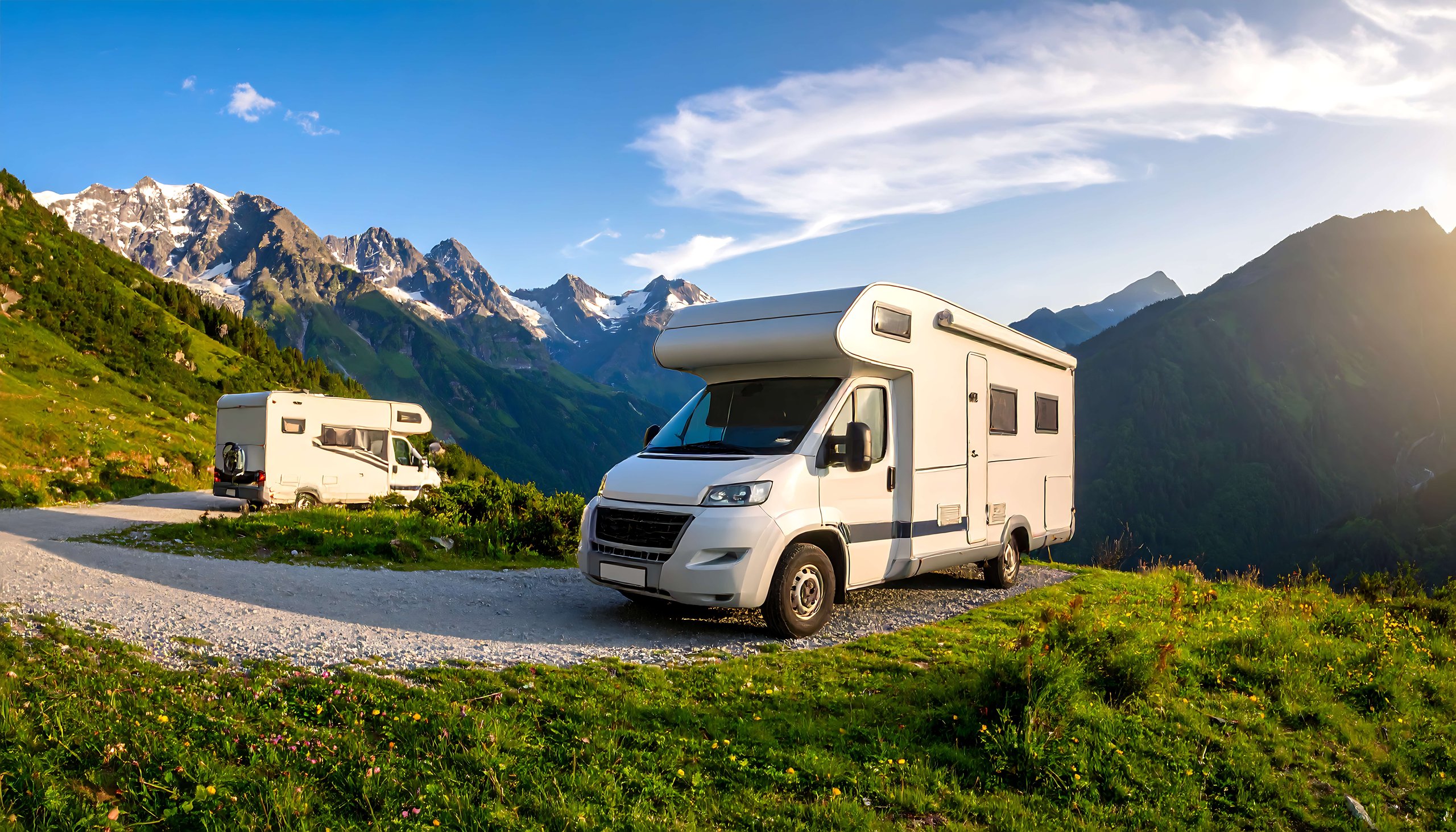 Zwei Wohnmobile auf einem Bergweg mit grünen Wiesen und schneebedeckten Bergen bei Sonnenuntergang.