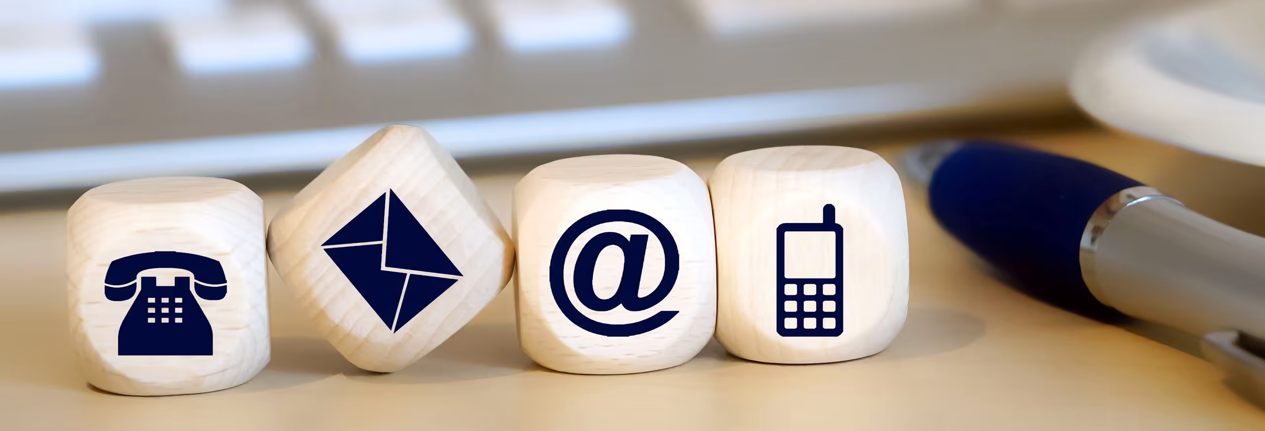 Vier Holzblöcke mit Symbolen für Festnetztelefon, Briefumschlag, E-Mail und Mobiltelefon nebeneinander auf einem Schreibtisch neben einem Kugelschreiber.