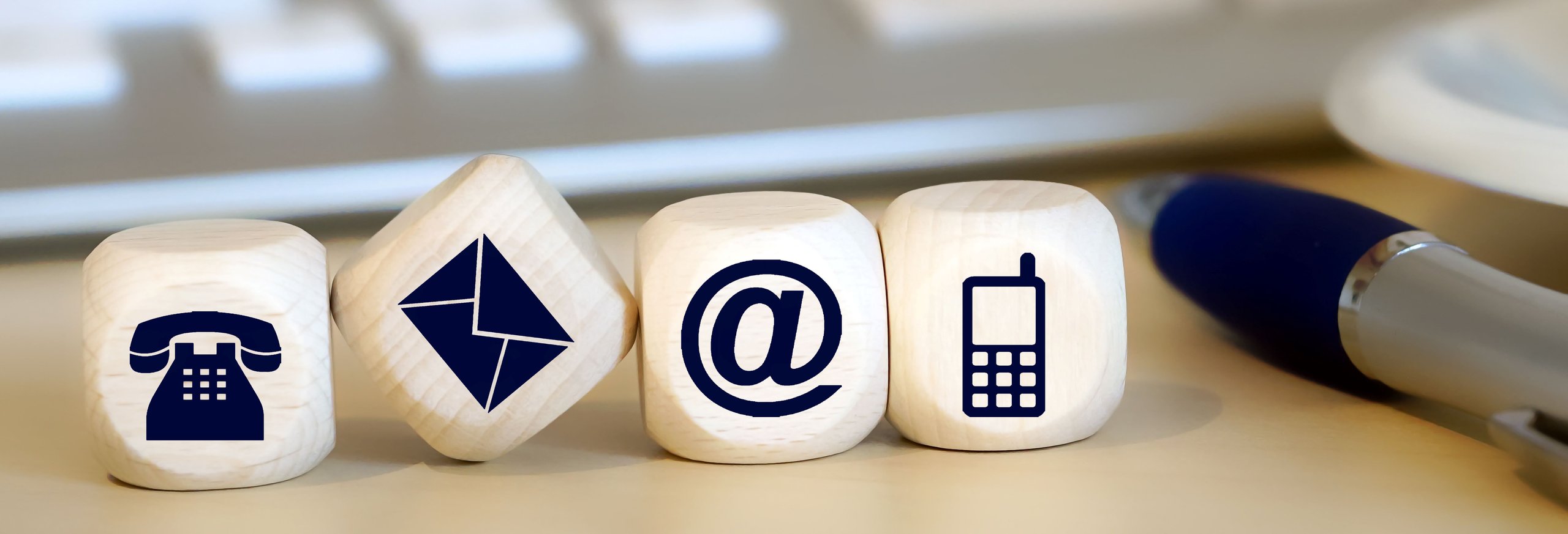 Vier Holzblöcke mit Symbolen für Festnetztelefon, Briefumschlag, E-Mail und Mobiltelefon nebeneinander auf einem Schreibtisch neben einem Kugelschreiber.
