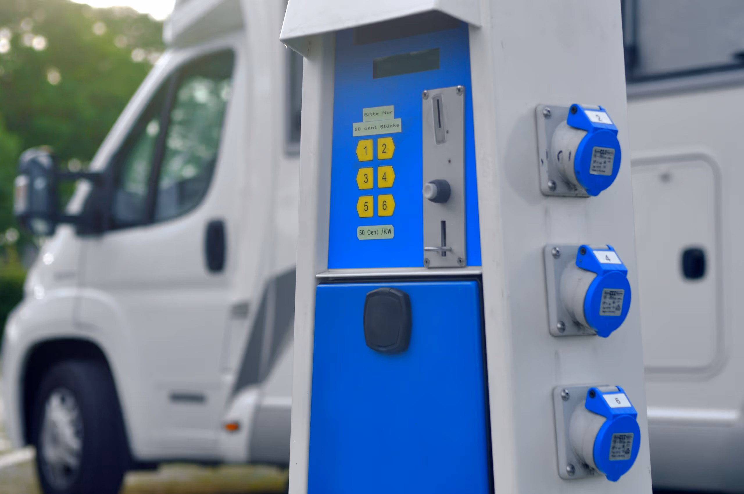 Elektrische Ladesäule mit Münzeinwurf und nummerierten Steckdosen neben einem weißen Wohnmobil.