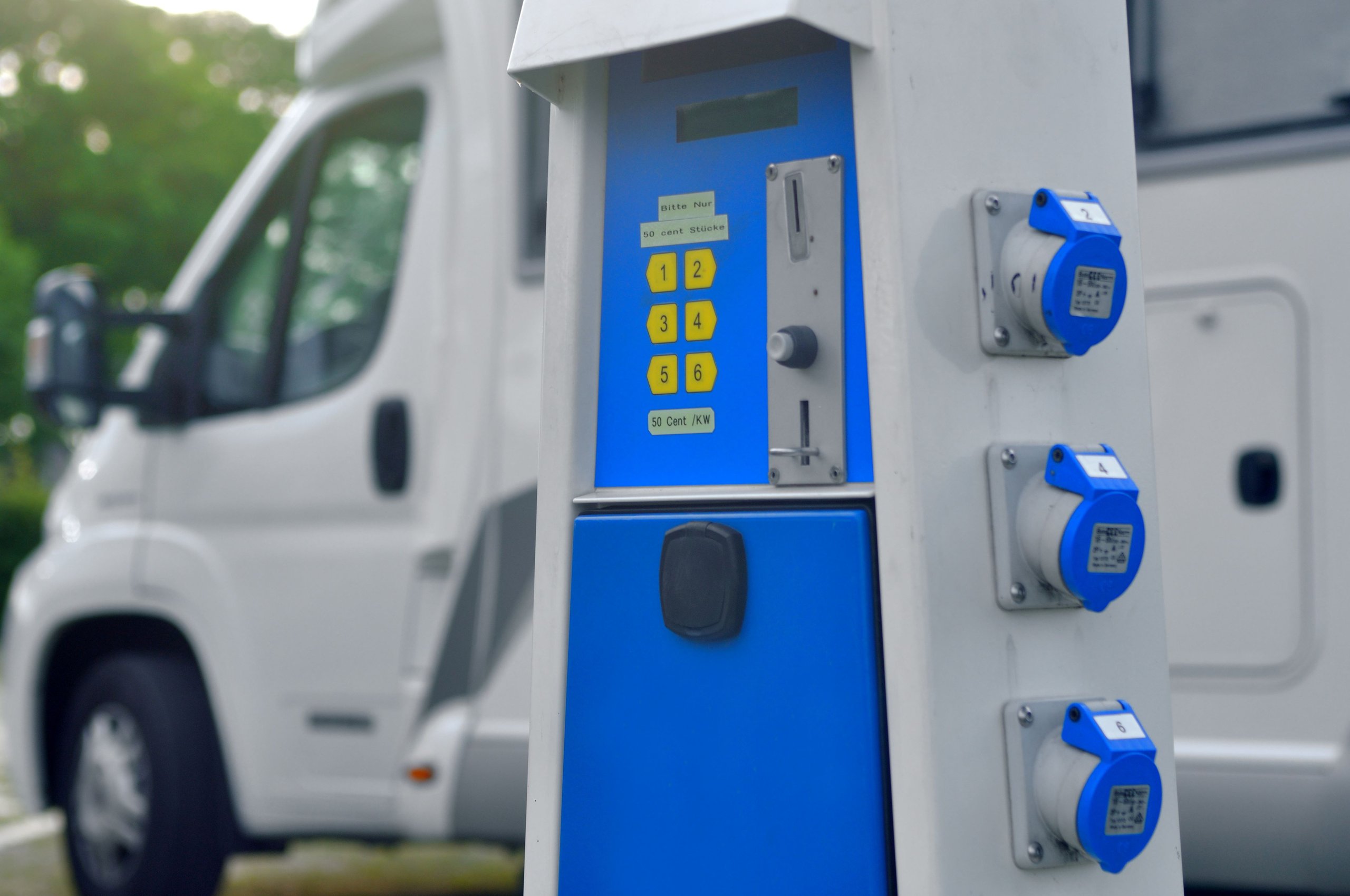 Elektrische Ladesäule mit Münzeinwurf und nummerierten Steckdosen neben einem weißen Wohnmobil.