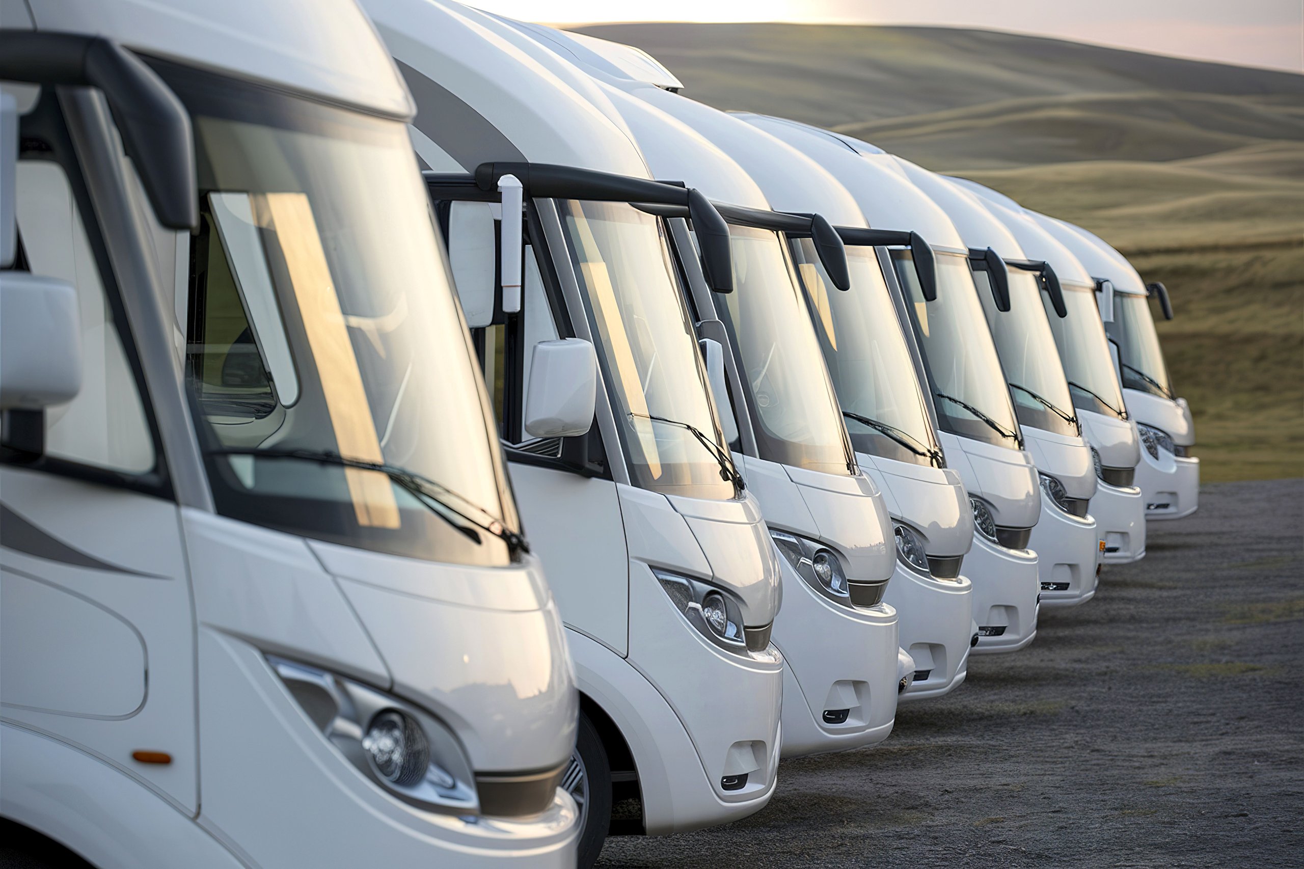 Mehrere weiße Wohnmobile stehen in einer Reihe auf einem offenen Gelände mit sanften Hügeln im Hintergrund.