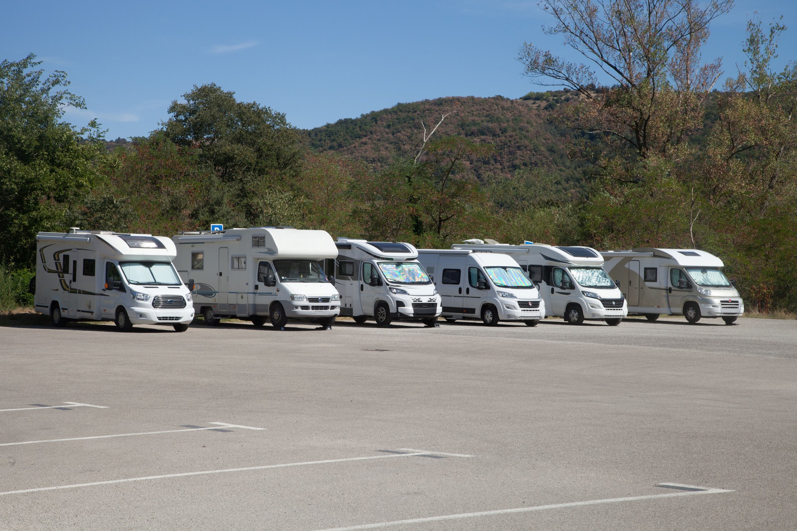 Sechs weisse Campervans auf einem Parkplatz mit bewaldeten Hügeln im Hintergrund unter blauem Himmel.