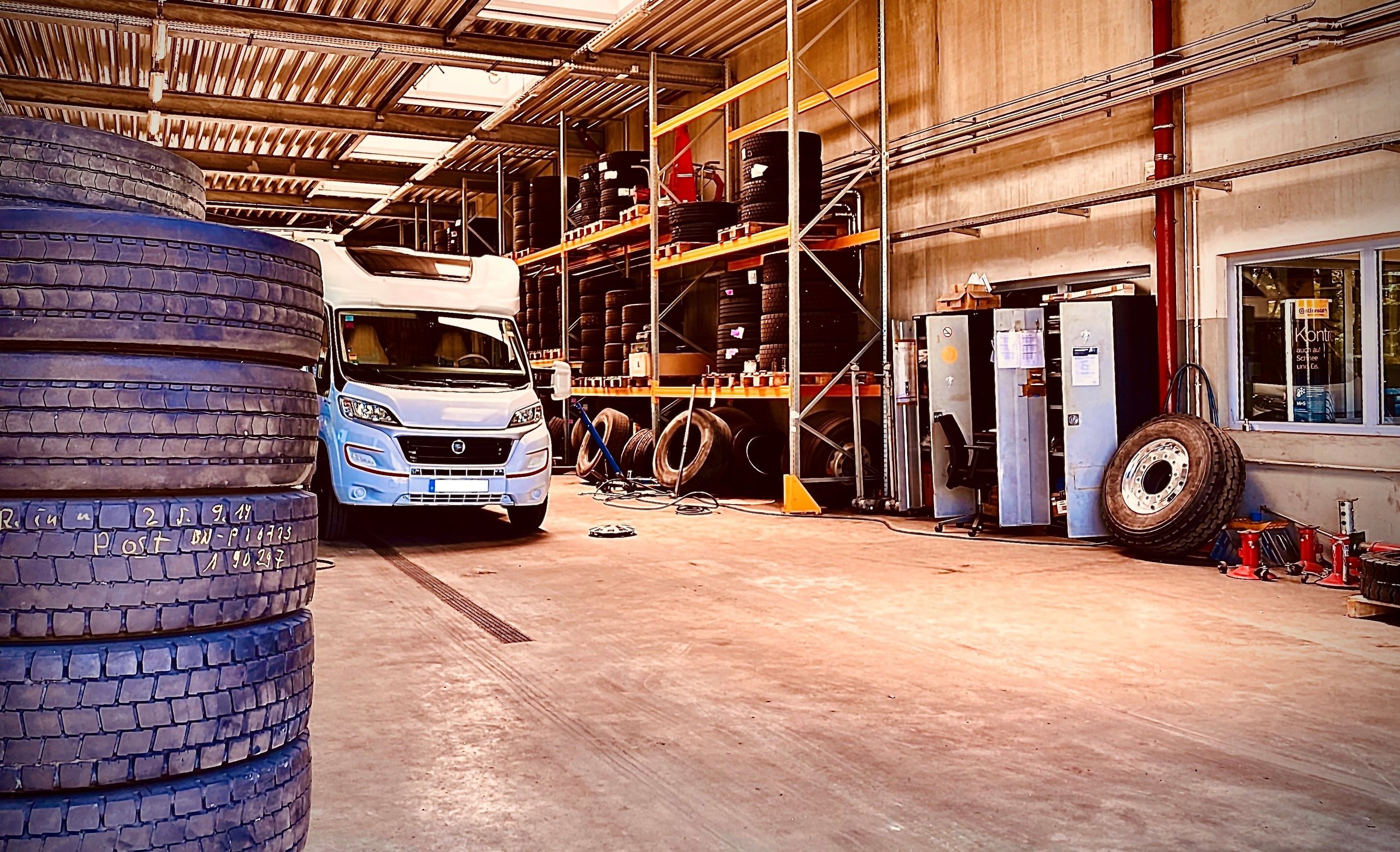 Weisse Nutzfahrzeugkabine in einer Lagerhalle mit gestapelten Reifen links und Regalen voller Reifen im Hintergrund.