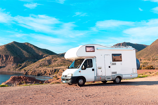 Weisser Camper parkiert auf einem kiesigen Ufer mit Bergen und blauem Himmel im Hintergrund.