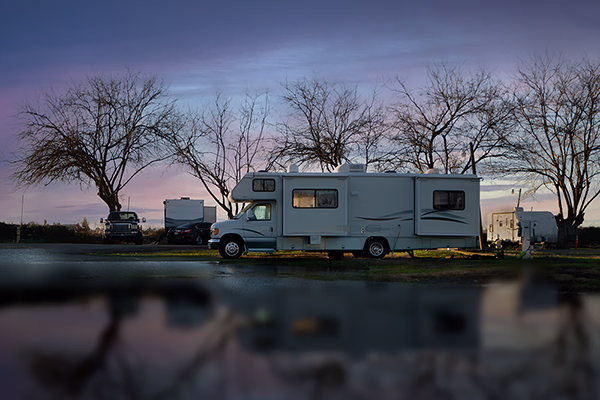Weisser Camper geparkt bei Dämmerung neben kahlen Bäumen mit Reflexion auf nasser Fläche im Vordergrund.