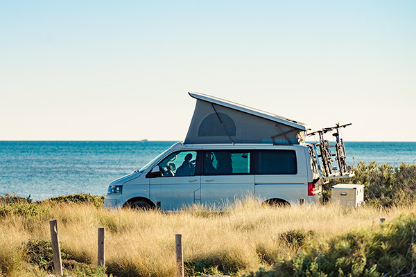 Weisser Camper-Van mit aufgeklapptem Dach und zwei Fahrrädern hinten, geparkt an einer Küste mit Gras und Meer im Hintergrund.