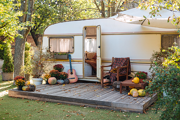 Gemütlicher Wohnwagen mit offener Tür, umgeben von Herbstdekorationen wie Kürbissen, Blumen und einer Gitarre auf einer Holzterrasse im Garten.