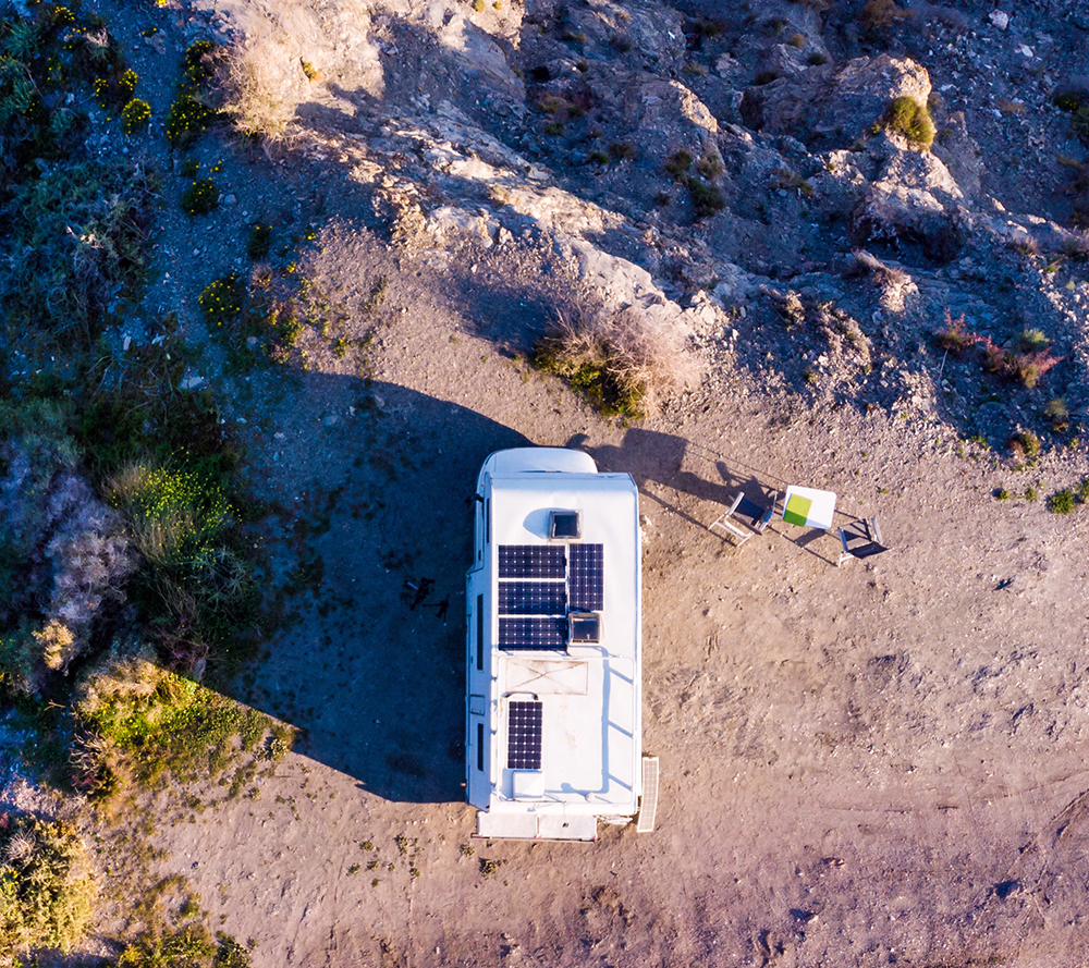 Weisses Camper mit Solarpanels auf dem Dach, daneben zwei Stühle und ein kleiner Tisch auf trockenem Boden.
