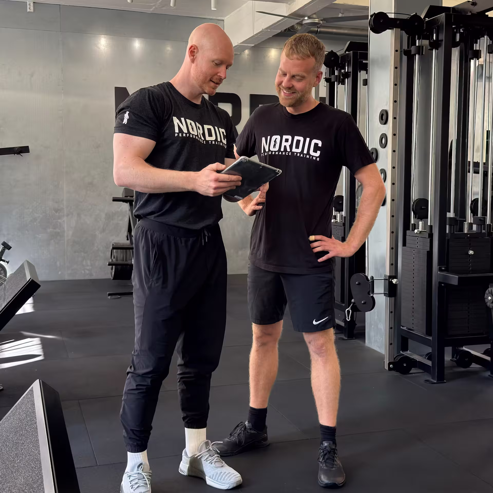 Mandlig klient smiler og er glad fordi hans personlige træner og fysioterapeut viser ham hans fremgang hen over ugerne på Ipad i Nordic Performance Training i København.
