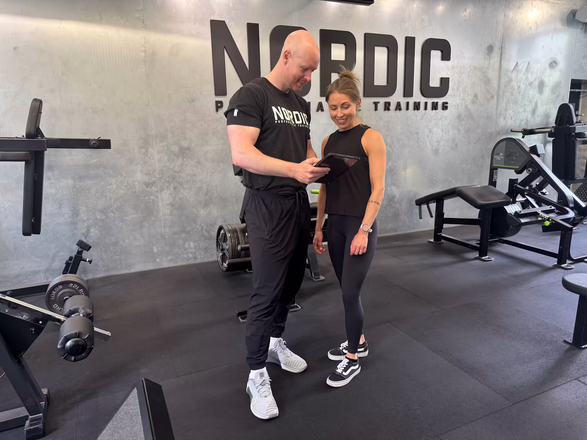 Personal trainer and physiotherapist at Nordic Performance Training’s private gym in Copenhagen showing a client her strength progress on an iPad, demonstrating visible results from just 1–2 sessions per week.