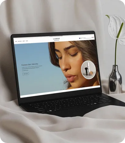 Laptop displaying the LAMAV website homepage with a close-up of a woman and skincare product, set against a soft fabric backdrop with a minimal vase decor.