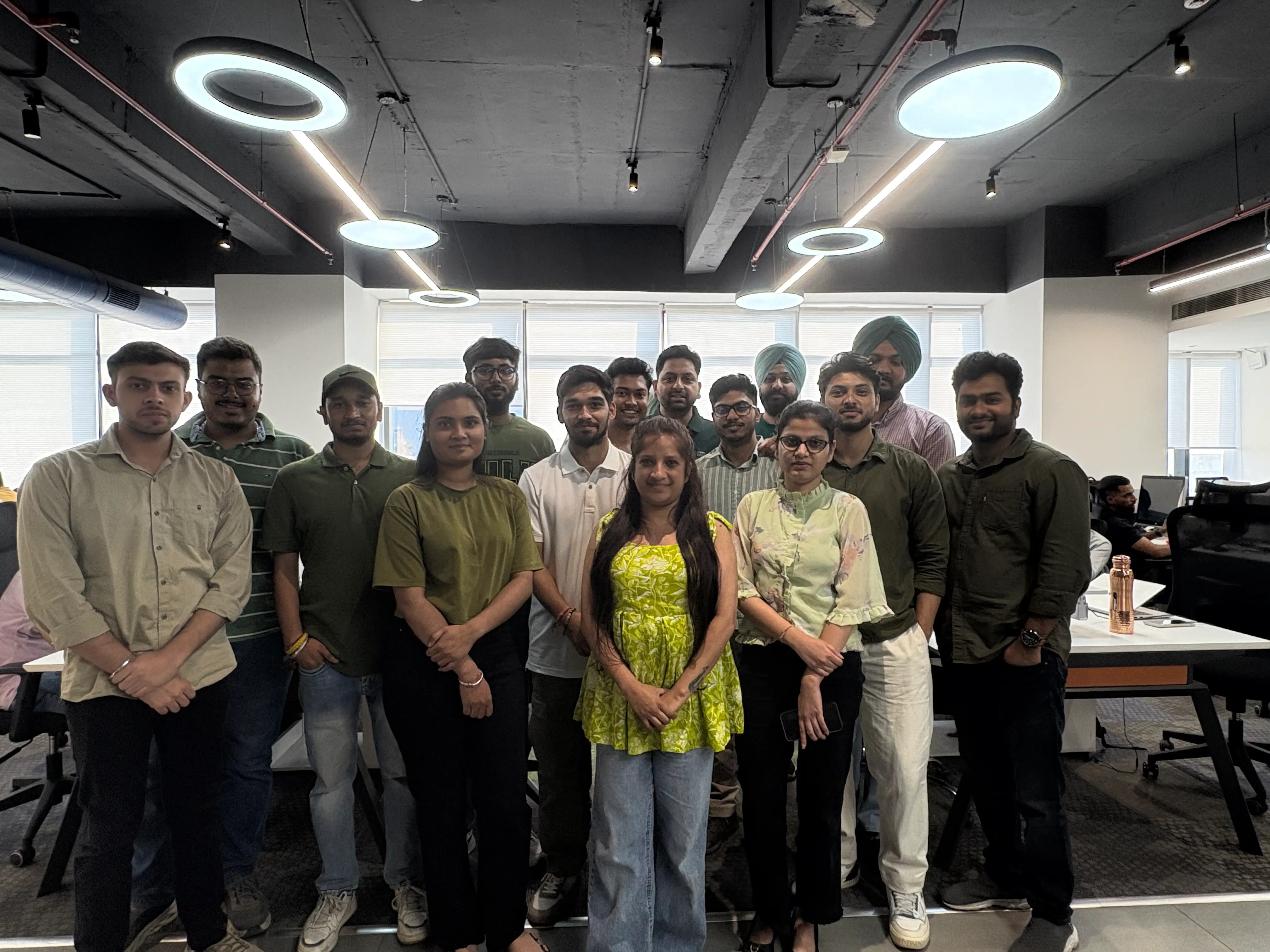 Group photo of team members dressed in shades of green for an office theme day, gathered together in a modern workspace under circular ceiling lights.
