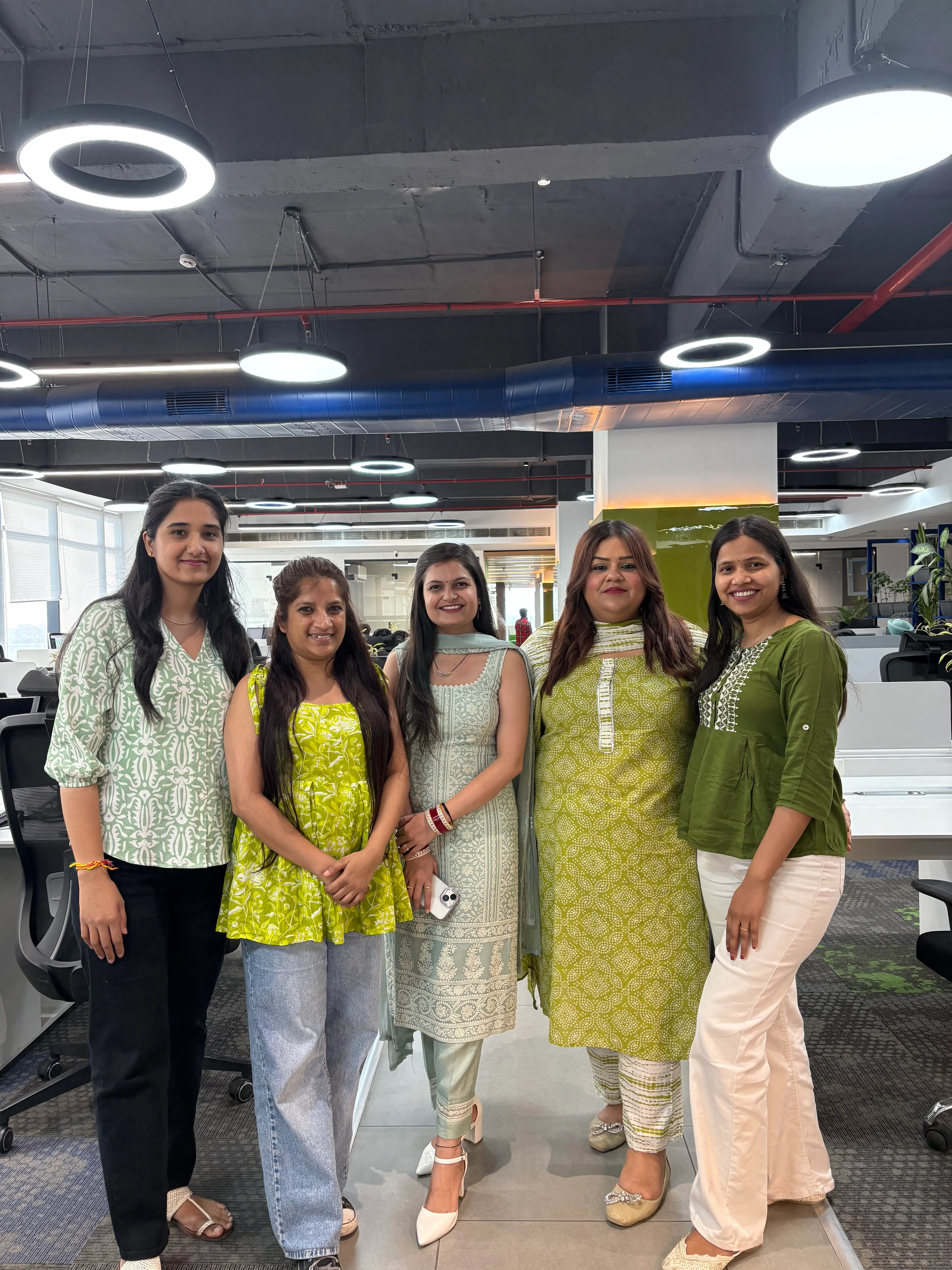 Five women team members dressed in coordinated green outfits posing together in a modern office space during a themed workday celebration.