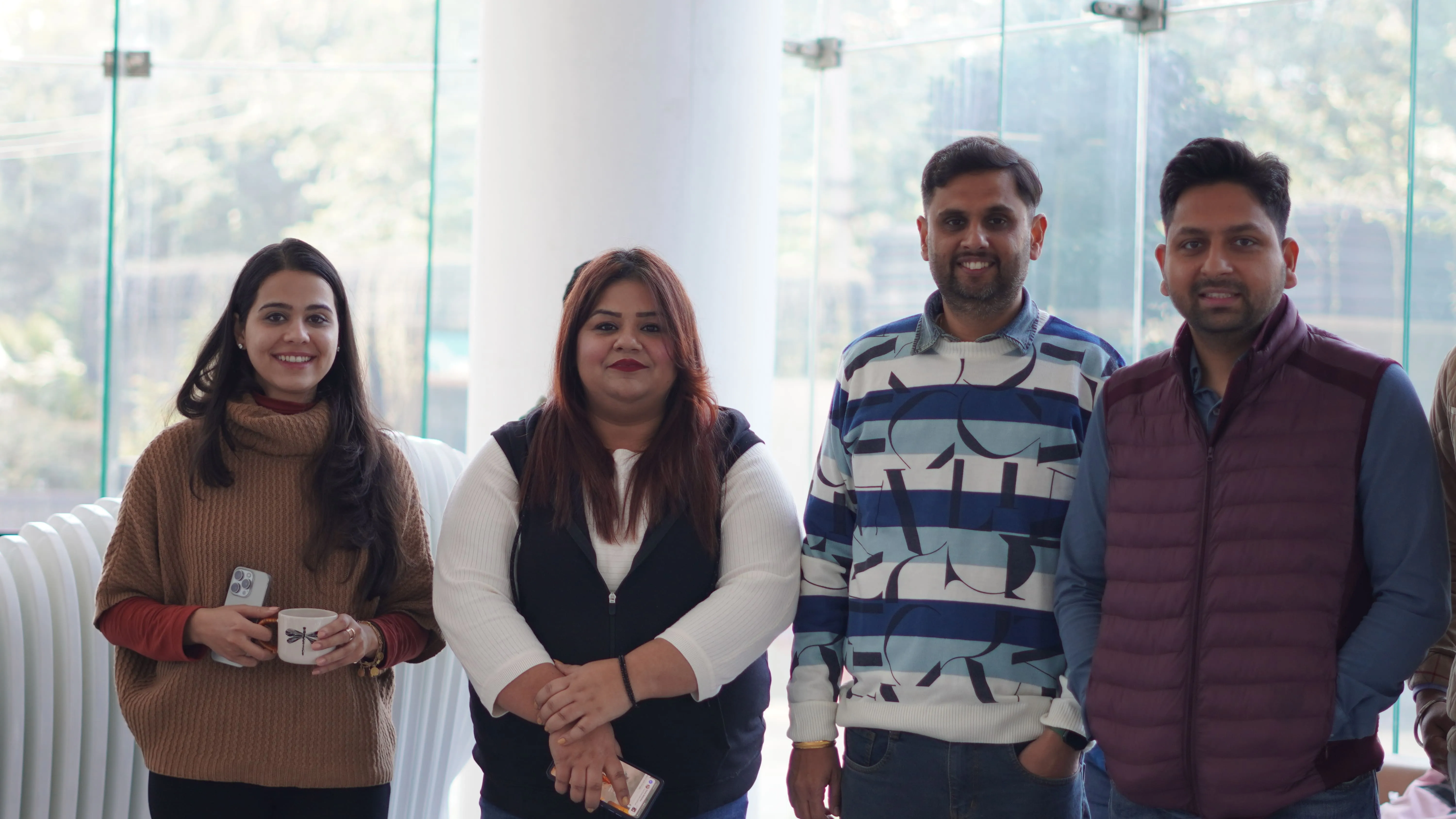 Group of four team members smiling and standing indoors in a well-lit office space, casually dressed and enjoying a relaxed moment together.