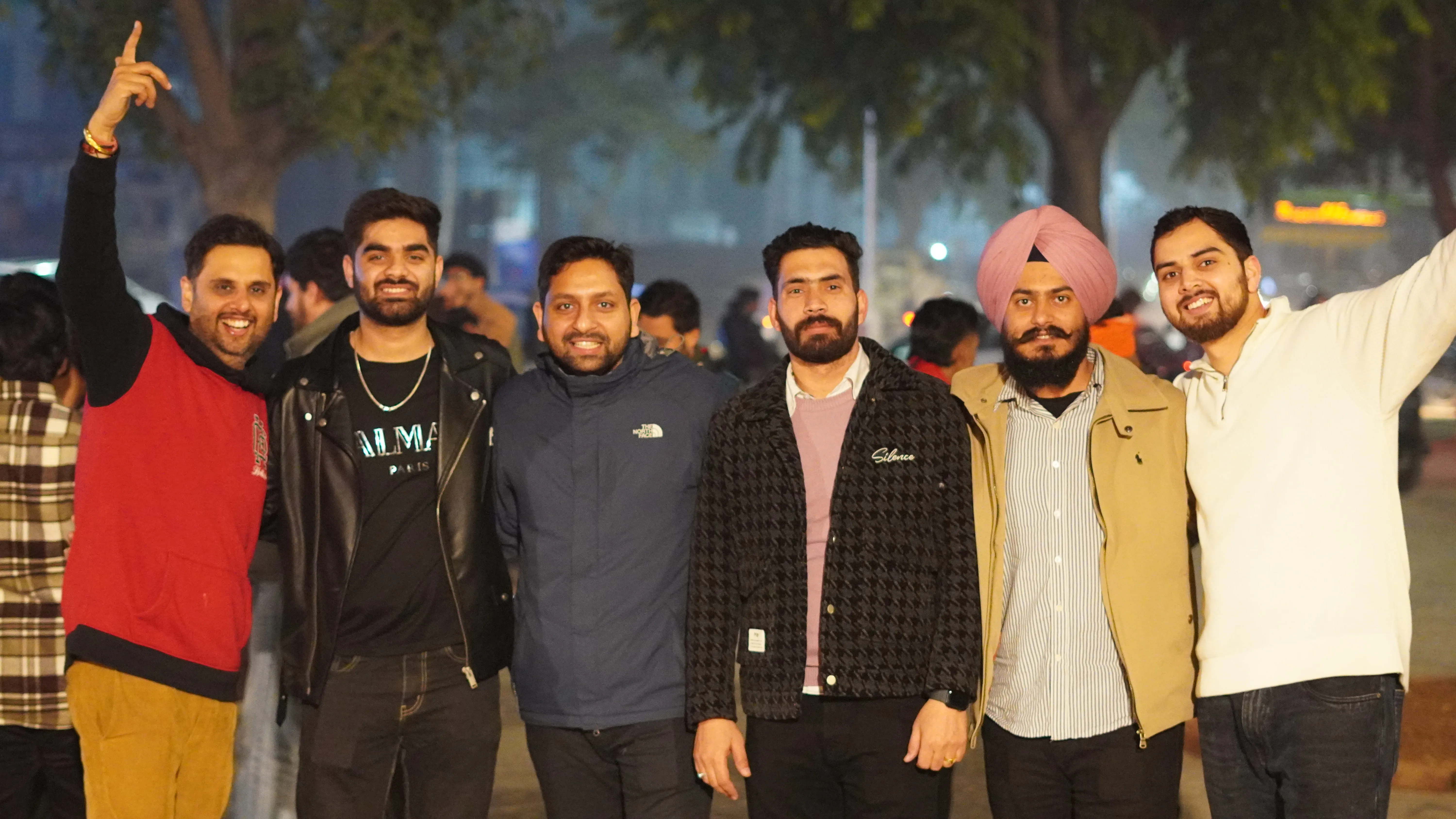 Group of six smiling men posing together outdoors during an evening event, with trees and lights in the background. Some of them have their arms around each other, showing camaraderie and celebration.