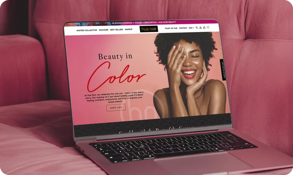 Laptop on a pink couch displaying the Hue Noir website with a smiling woman and “Beauty in Color” headline, promoting inclusive beauty products.