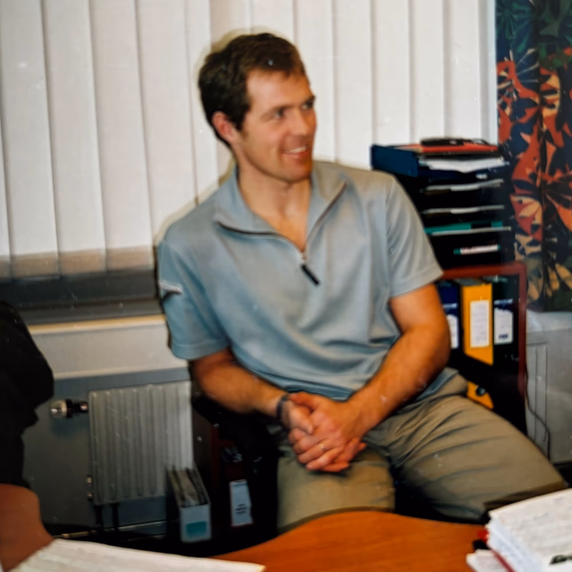 Man with short brown hair sitting at a desk, wearing a light gray short-sleeve shirt and beige pants, smiling and looking to the side.