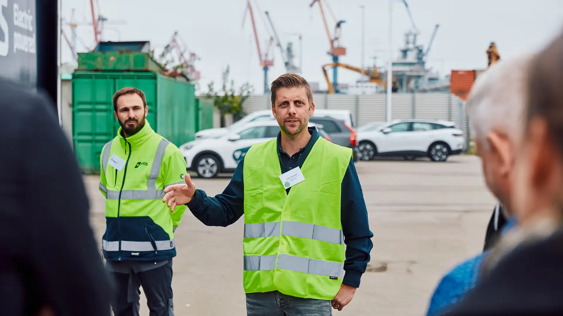 Two men wearing high-visibility vests speaking to an audience outdoors at an industrial site with cranes and parked cars in the background.