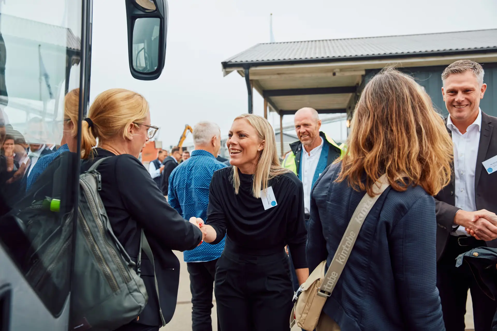 Two women shaking hands and smiling during an outdoor event with other people mingling in the background.