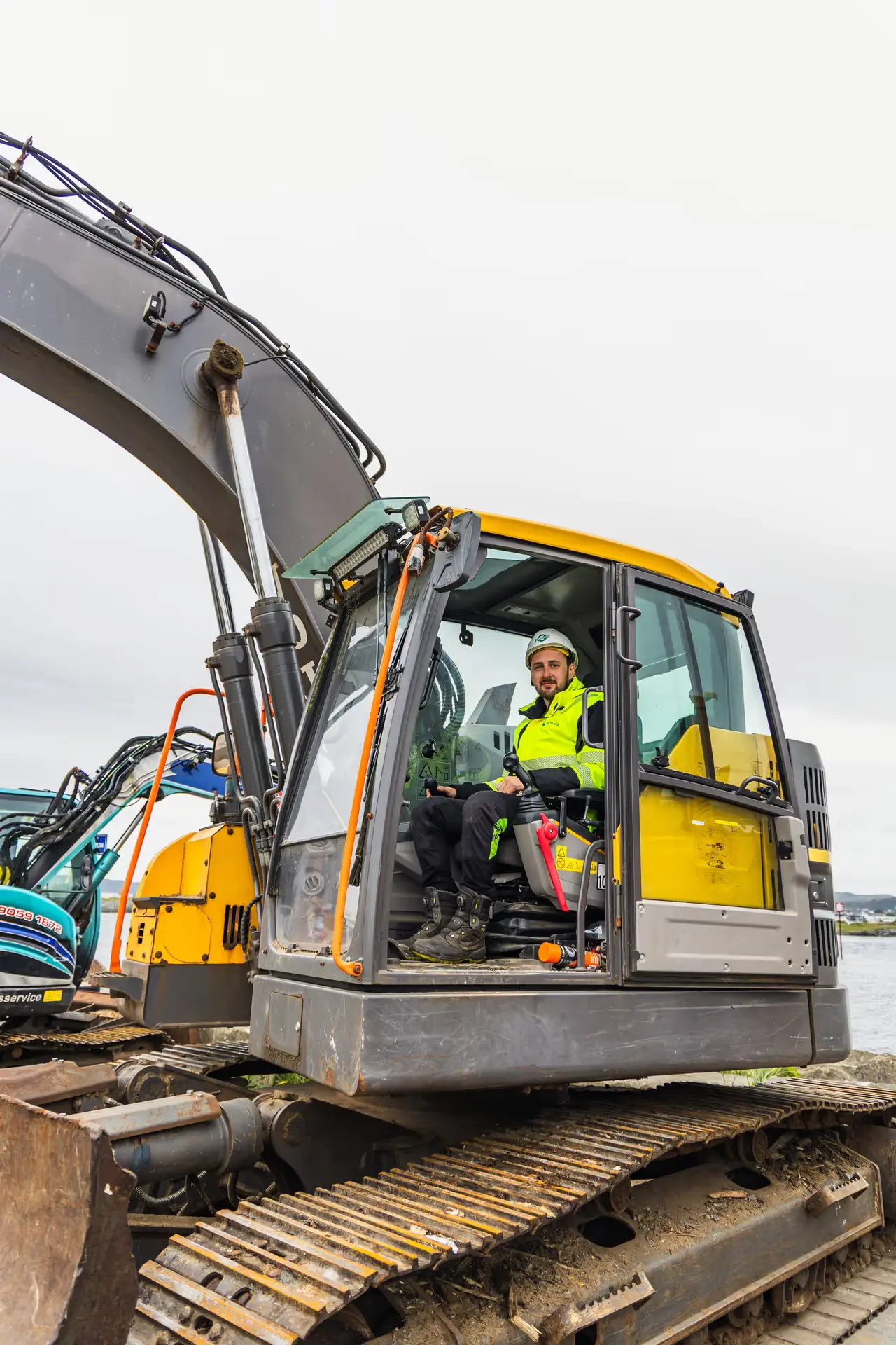 Construction worker wearing a hard hat and yellow safety jacket operating a yellow and gray excavator.