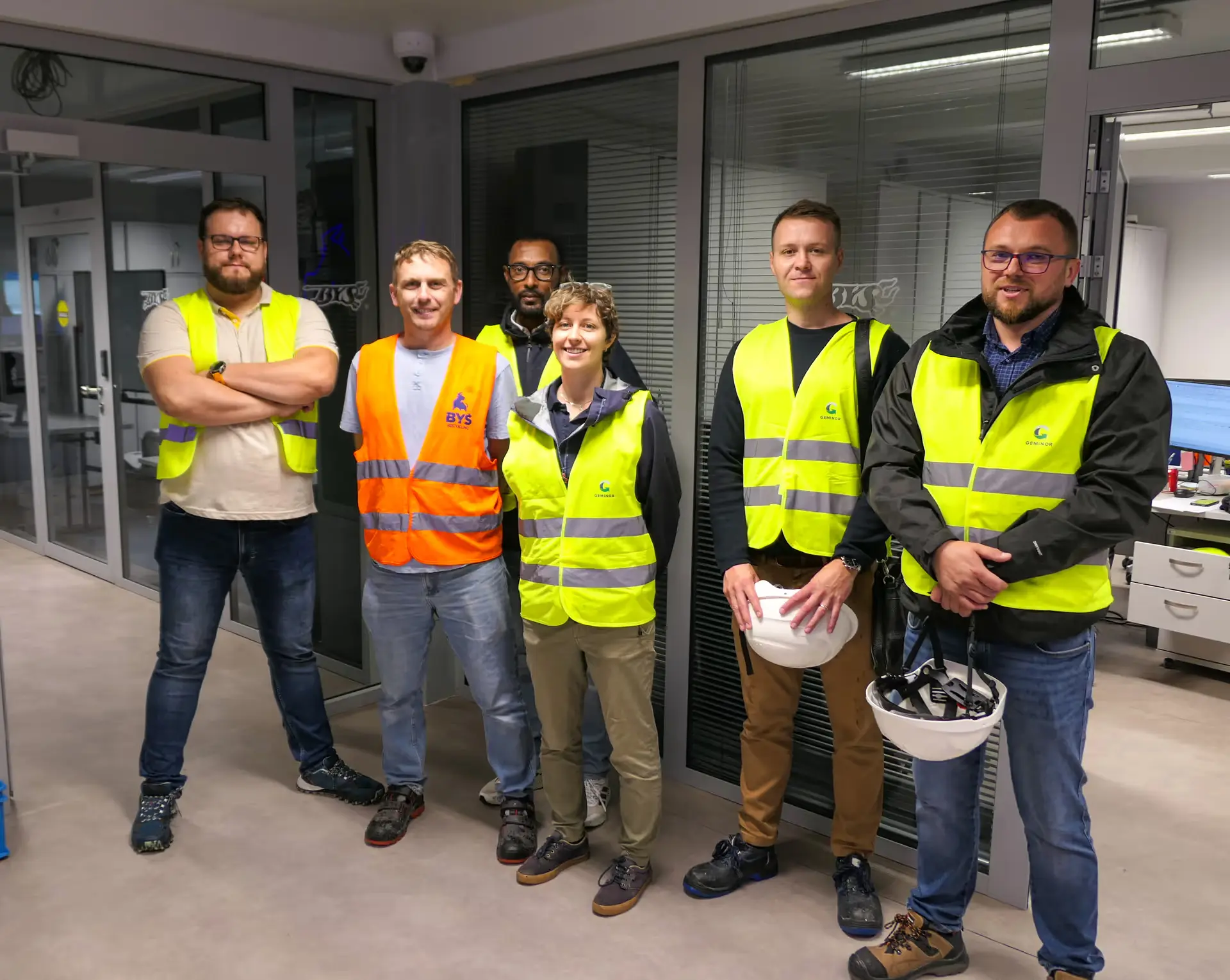 Group of six construction workers wearing safety vests, two holding white hard hats, standing indoors in an office-like environment.