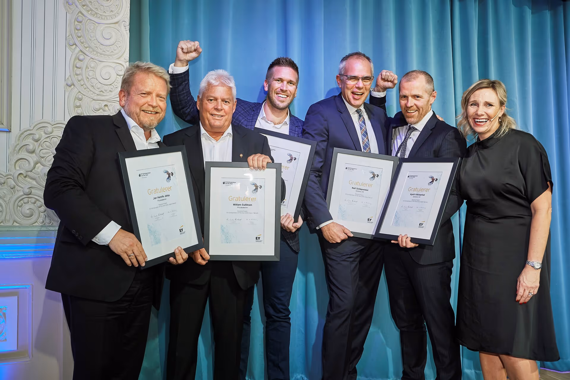 Six people in formal attire smiling and holding framed certificates in front of a blue curtain, with two men raising their fists in celebration.