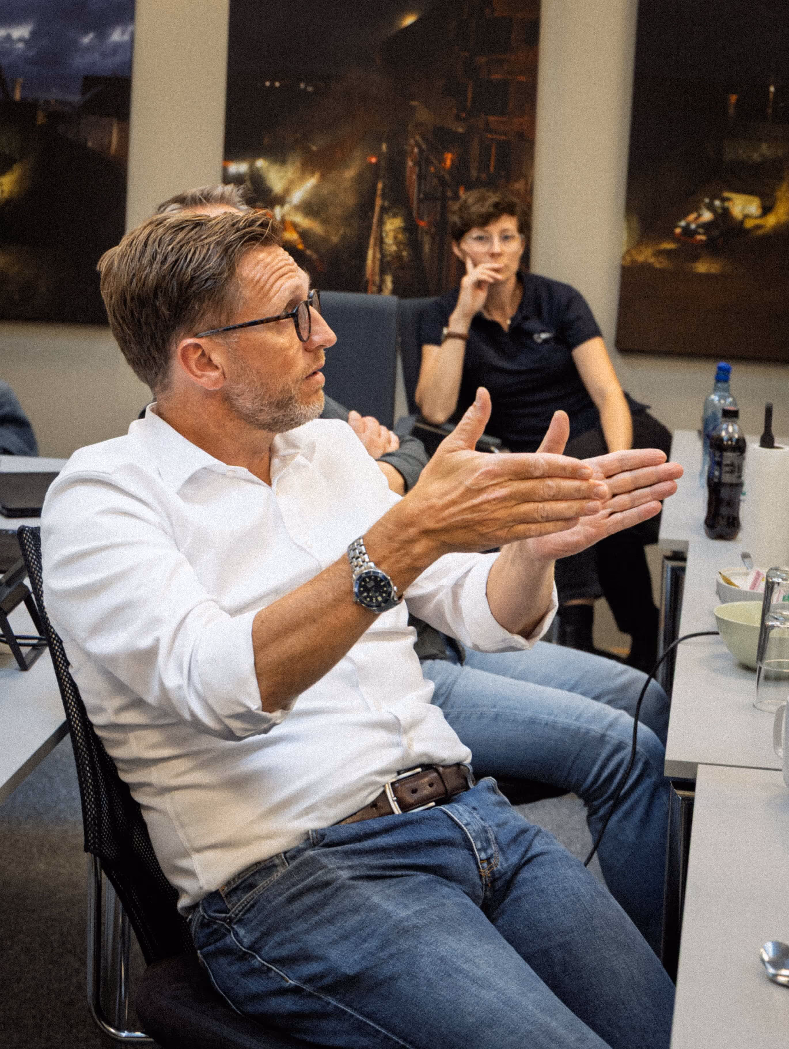 Man in white shirt and glasses gesturing with his hands while seated at a meeting table with other attentive people in the background.