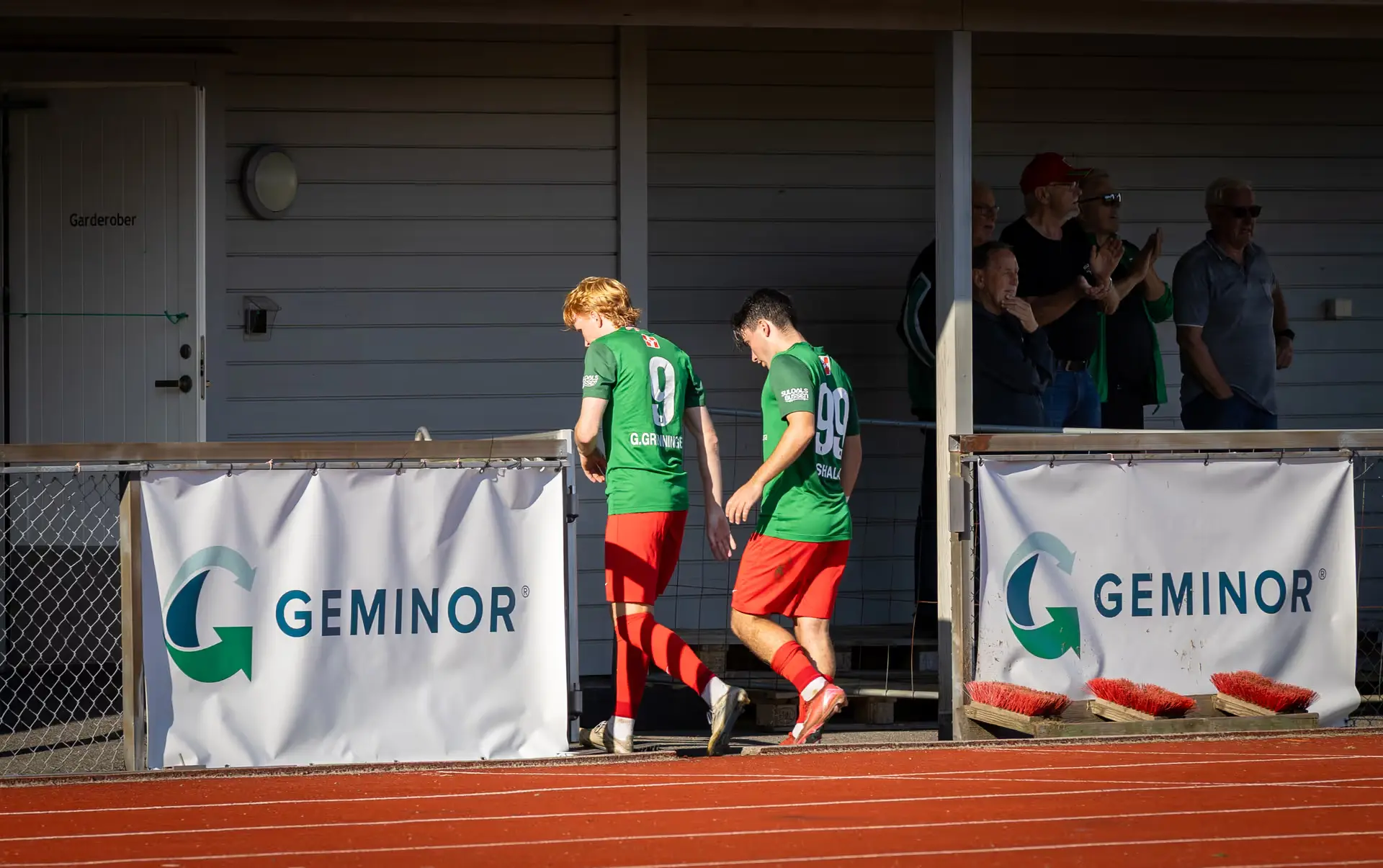 Two soccer players in green jerseys and red shorts walking on a track near a wall with GEMINOR banners and spectators clapping.