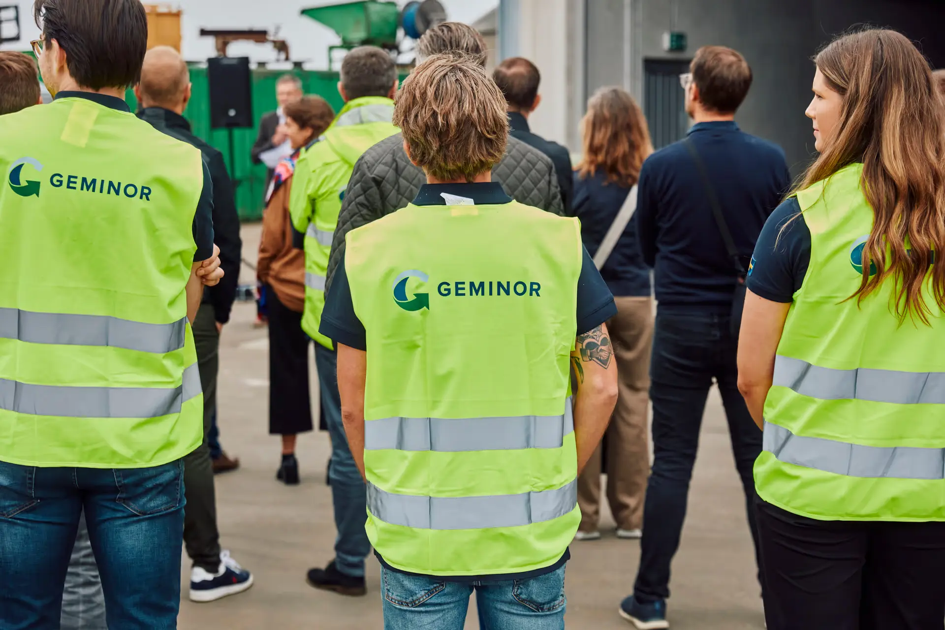 Group of people standing indoors, three wearing high-visibility vests labeled GEMINOR, facing away from the camera.