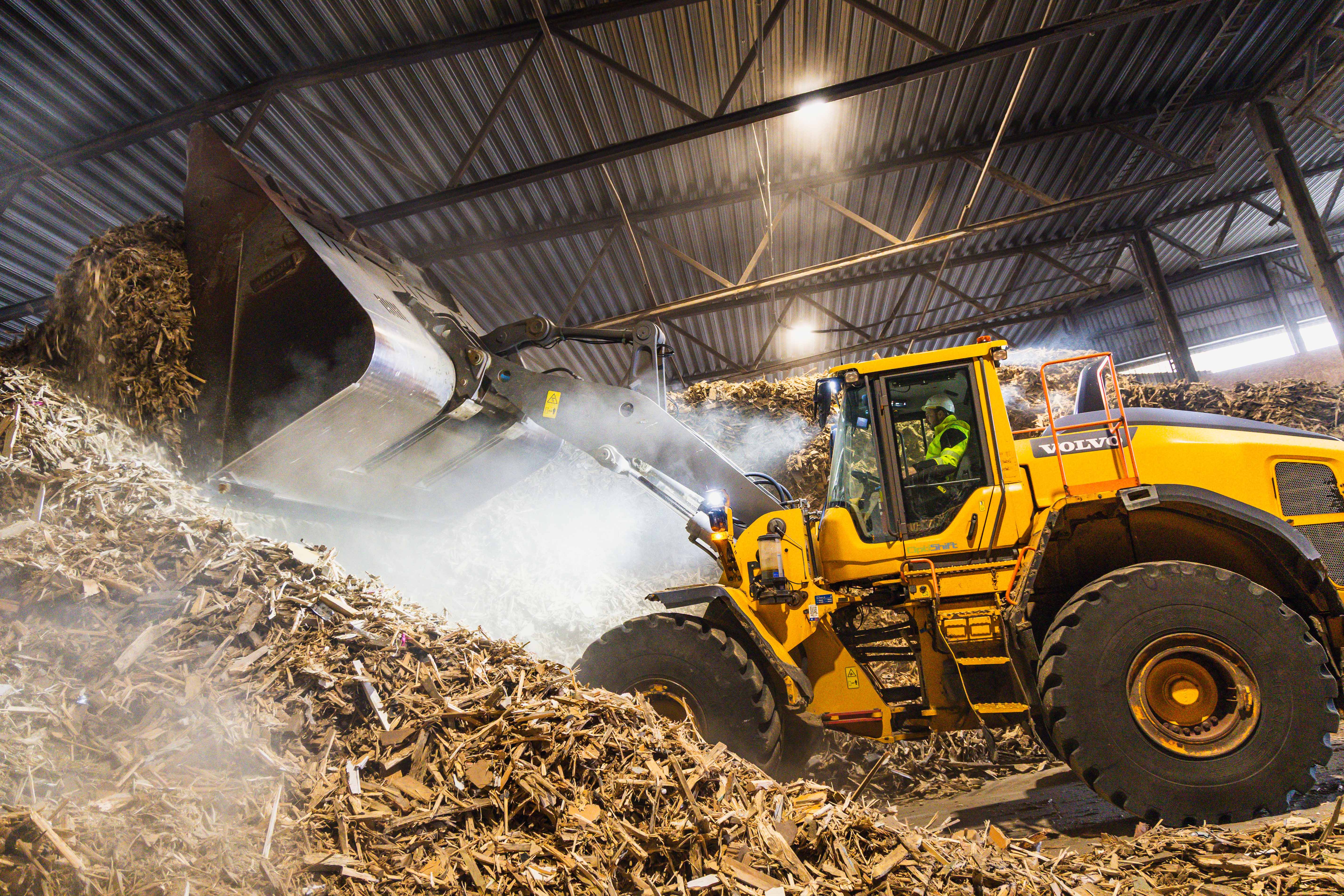 Yellow Volvo wheel loader moving a large pile of wood chips inside an industrial facility.