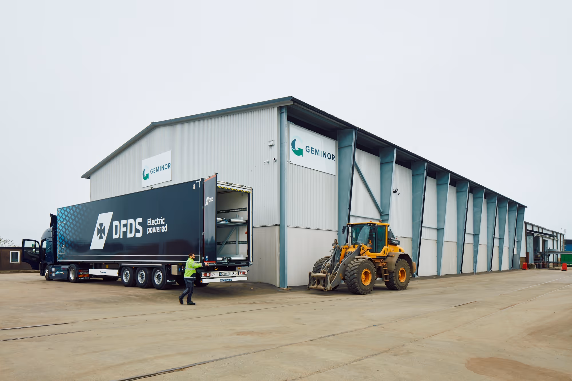 A DFDS electric-powered truck backed up to a large warehouse with GEMINOR signs, next to a yellow construction loader and a person in a green jacket.