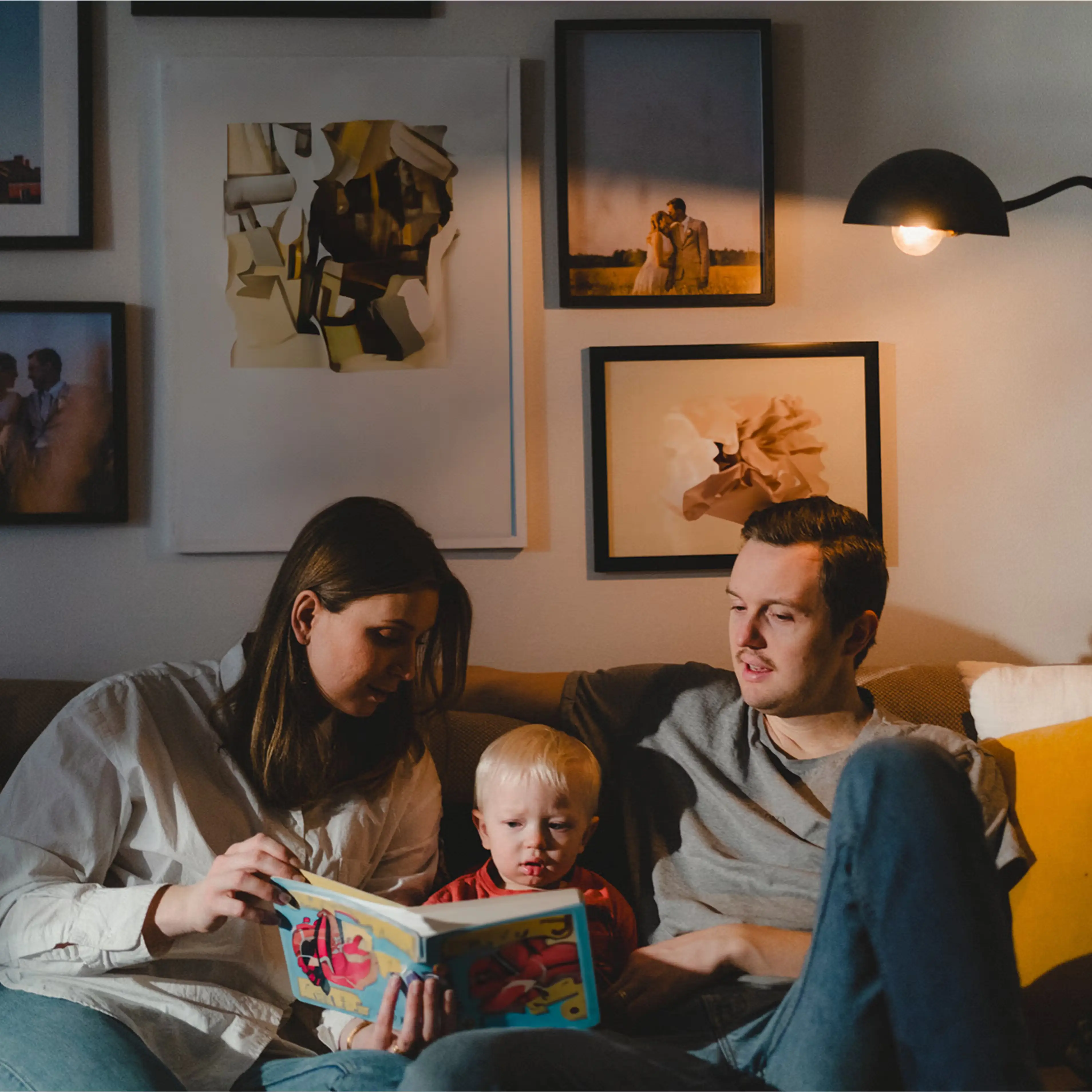 A mother and father sitting on a couch reading a children's book with their young child between them.