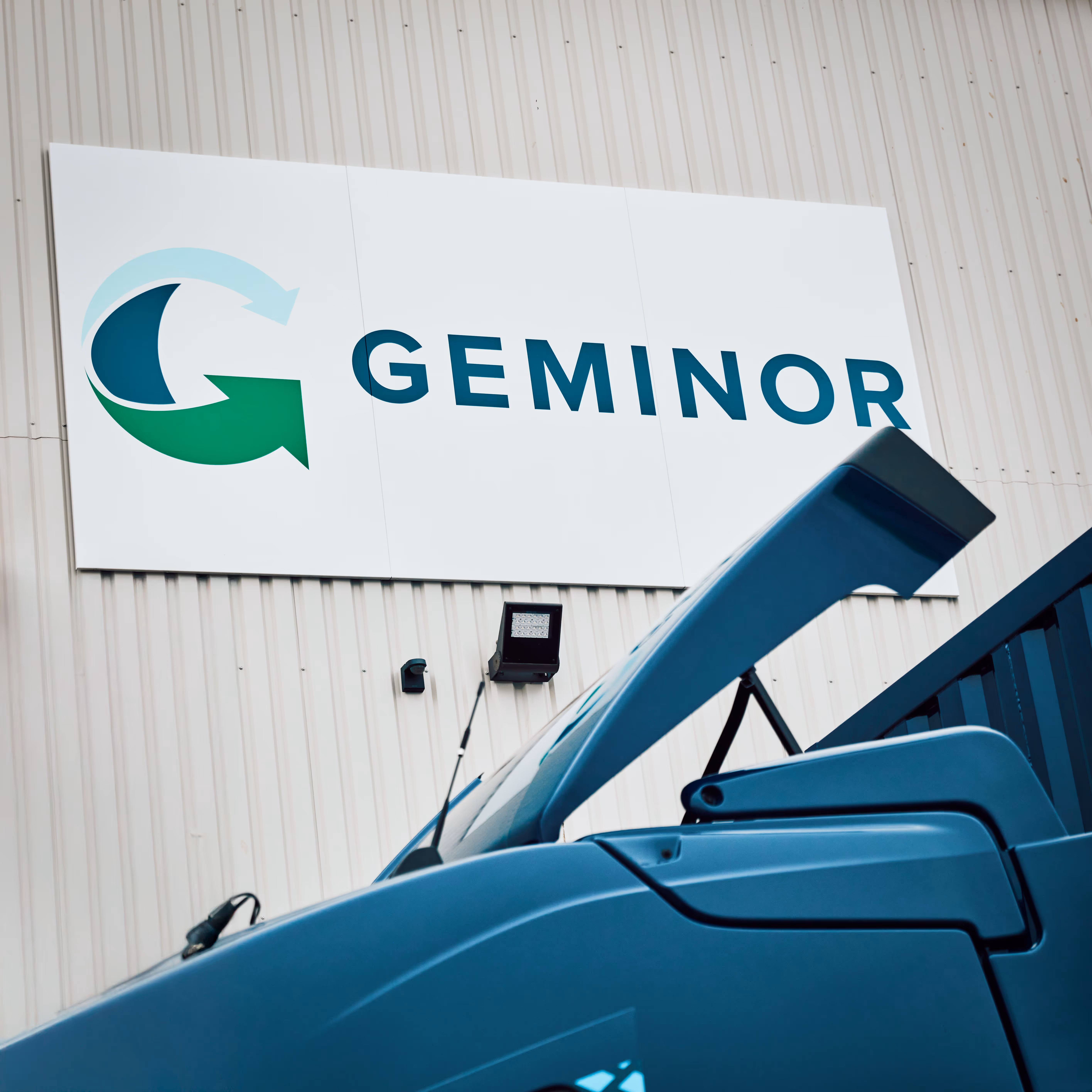 Close-up of a blue truck with an open door in front of a building featuring a large Geminor company logo sign.