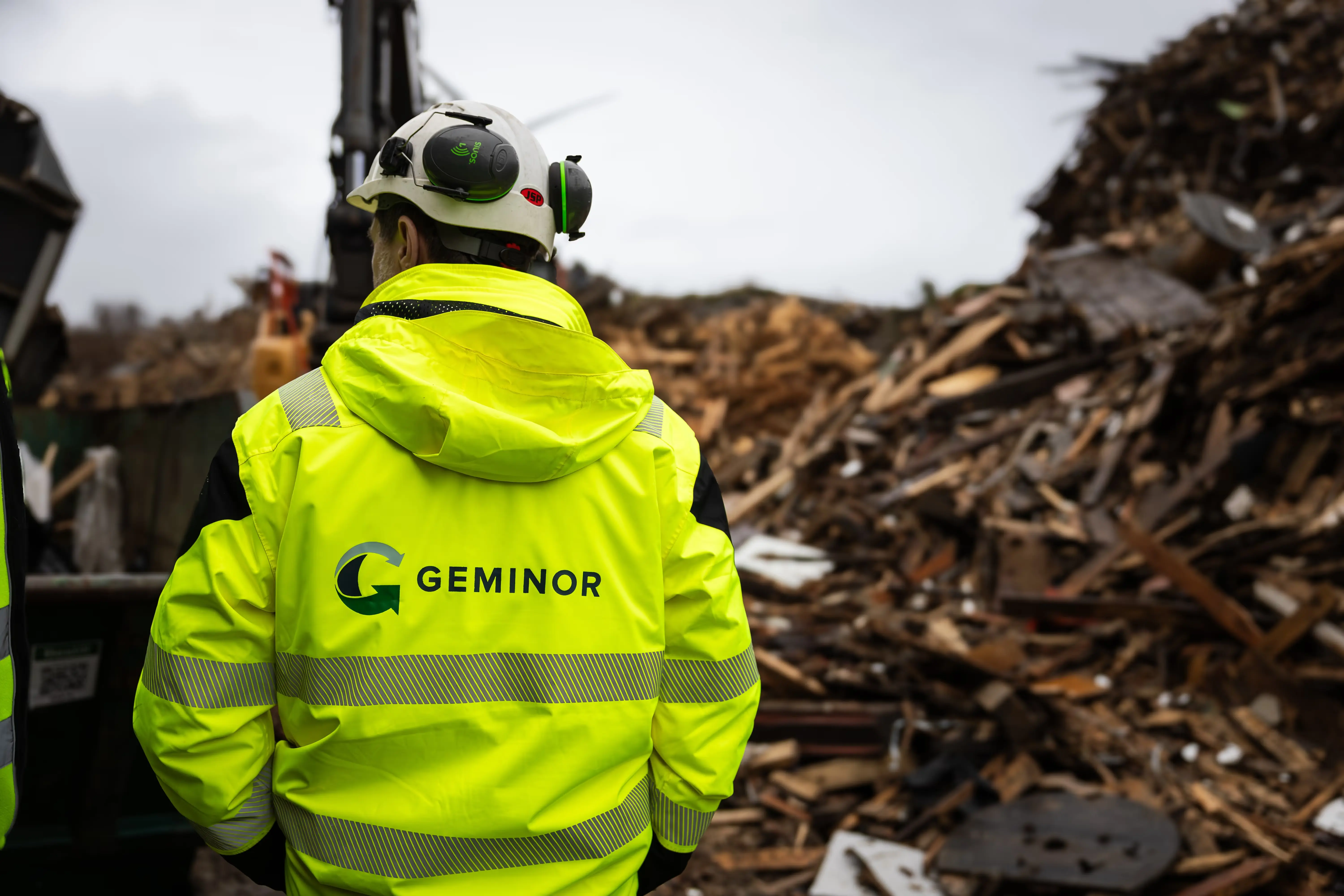 Two workers in yellow high-visibility jackets and white helmets inspecting a large pile of wood debris at a construction or demolition site.
