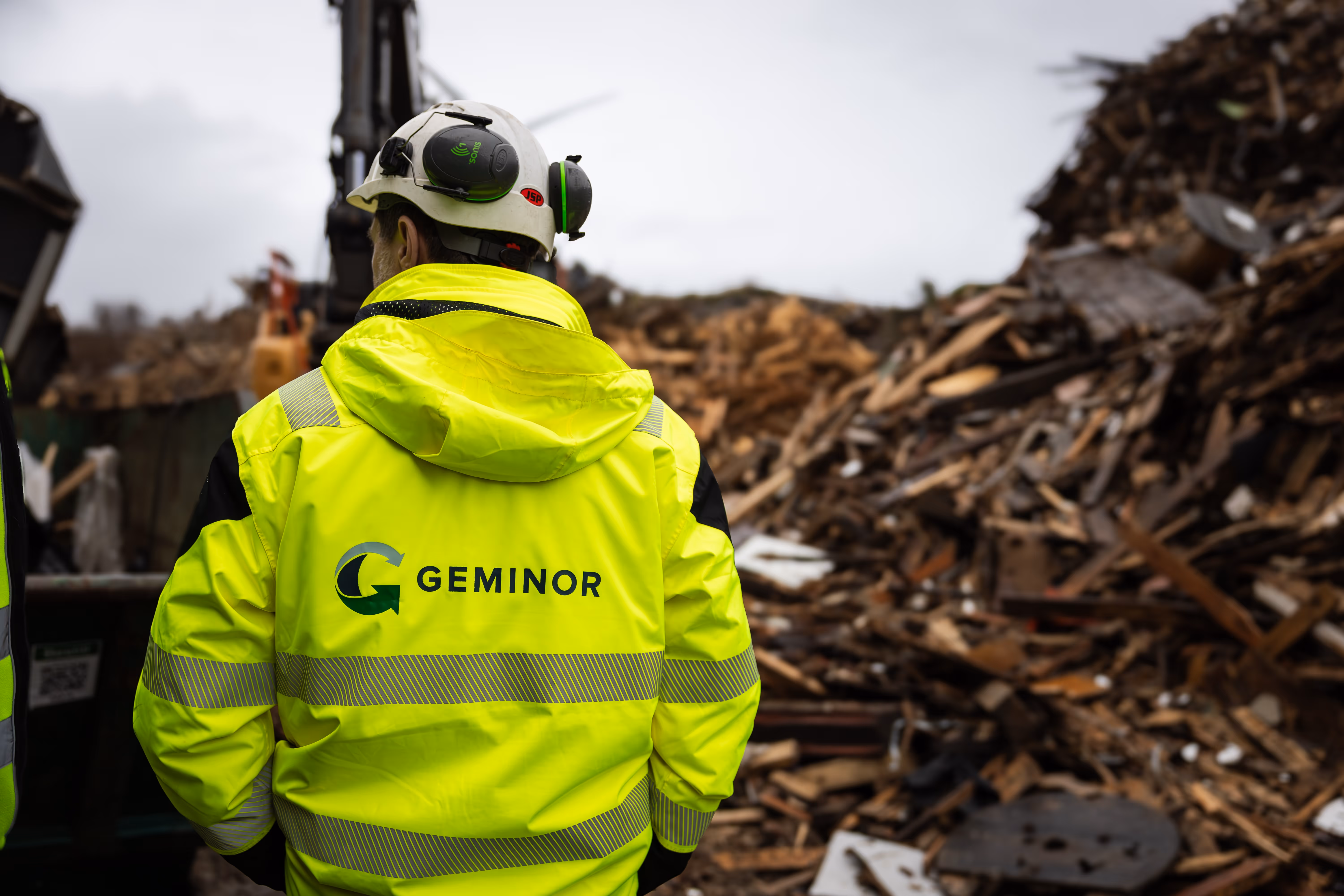Two workers in yellow high-visibility jackets and white helmets inspecting a large pile of wood debris at a construction or demolition site.