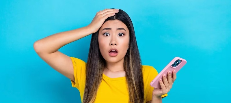 Teenage girl in a yellow shirt holding a pink smartphone, looking shocked with one hand on her head, standing in front of a bright blue background. The image conveys surprise, alarm, or digital distress—ideal for illustrating online safety, cyberbullying, or social media risks.