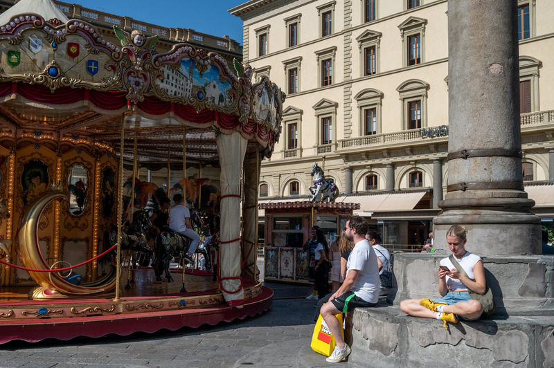 People relaxing near the historic carousel at Piazza della Repubblica in Florence, Italy. A woman sits cross-legged using her phone, while a man with a LEGO bag looks on. The carousel and classic architecture in the background highlight the blend of tradition and modern life in this popular tourist spot.