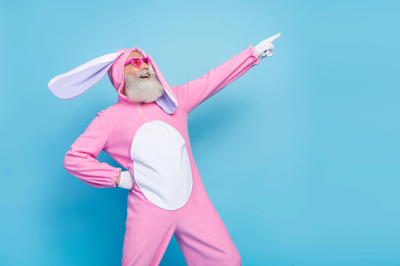 Elderly man with a white beard wearing a playful pink bunny costume and heart-shaped sunglasses, confidently pointing upward with a joyful expression against a bright blue background, celebrating humor, self-expression, and ageless fun.