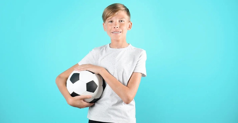 Teenage boy with braces and a light t-shirt smiling while holding a black and white soccer ball against a bright blue background, representing youth sports, physical activity, and healthy lifestyle choices.