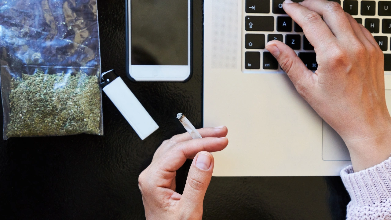 Homme qui tappe sur son ordinateur avec un joint à la main et un sachet de fleurs de CBD