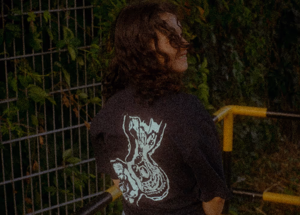 Renée Customs T-Shirt vor Metallzaun und grüner Bepflanzung – Produktfoto