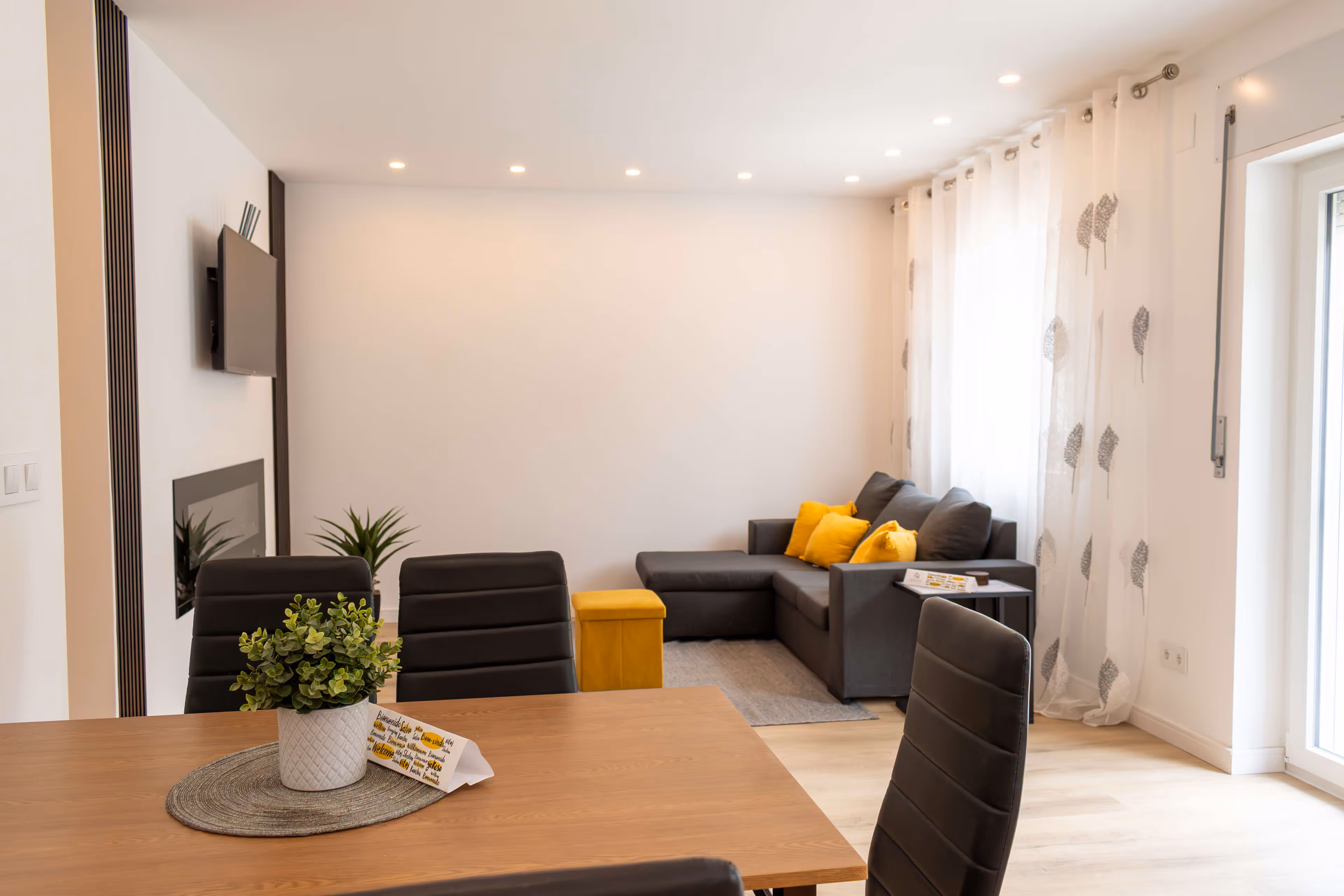 Modern living room with gray sectional sofa, yellow cushions, a wooden dining table with black chairs, and a potted plant centerpiece.
